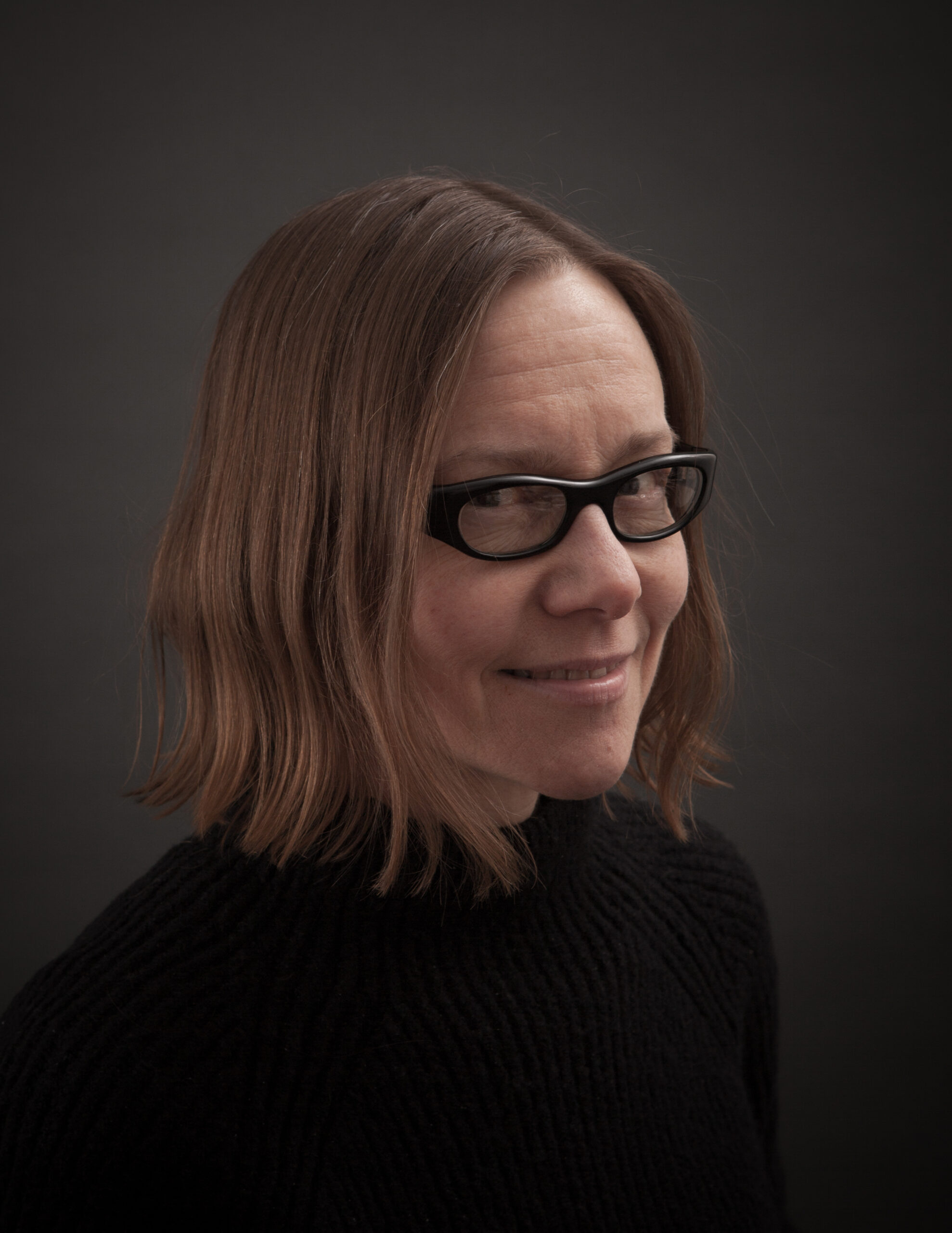 Cathie Axe. Image of a woman with shoulder length hair and black framed glasses against a dark background and in a black shirt.