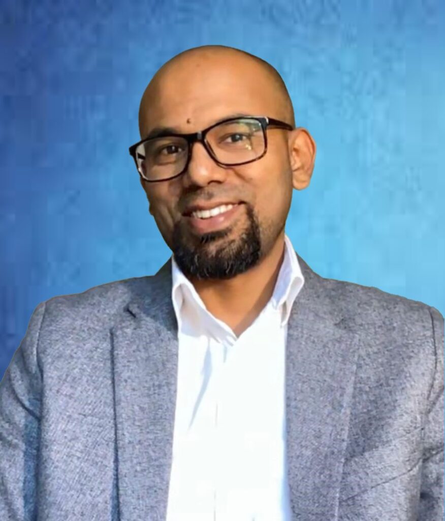 Syed Manzoor Ahmed. Image of a man wearing black framed glasses, a white collared shirt and light grey jacket against a blue background.