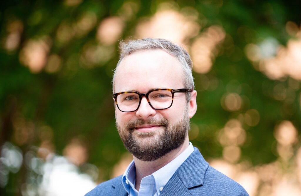 An image of a man in dark rimmed glasses and a blue suite jacket.