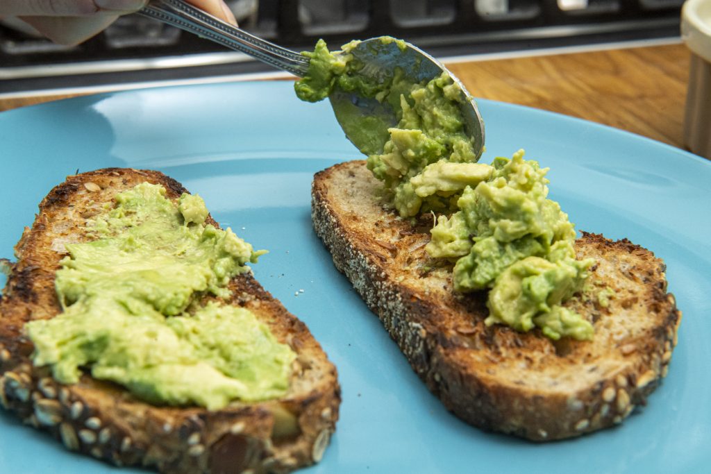 A spoon placing mashed avocado on toast