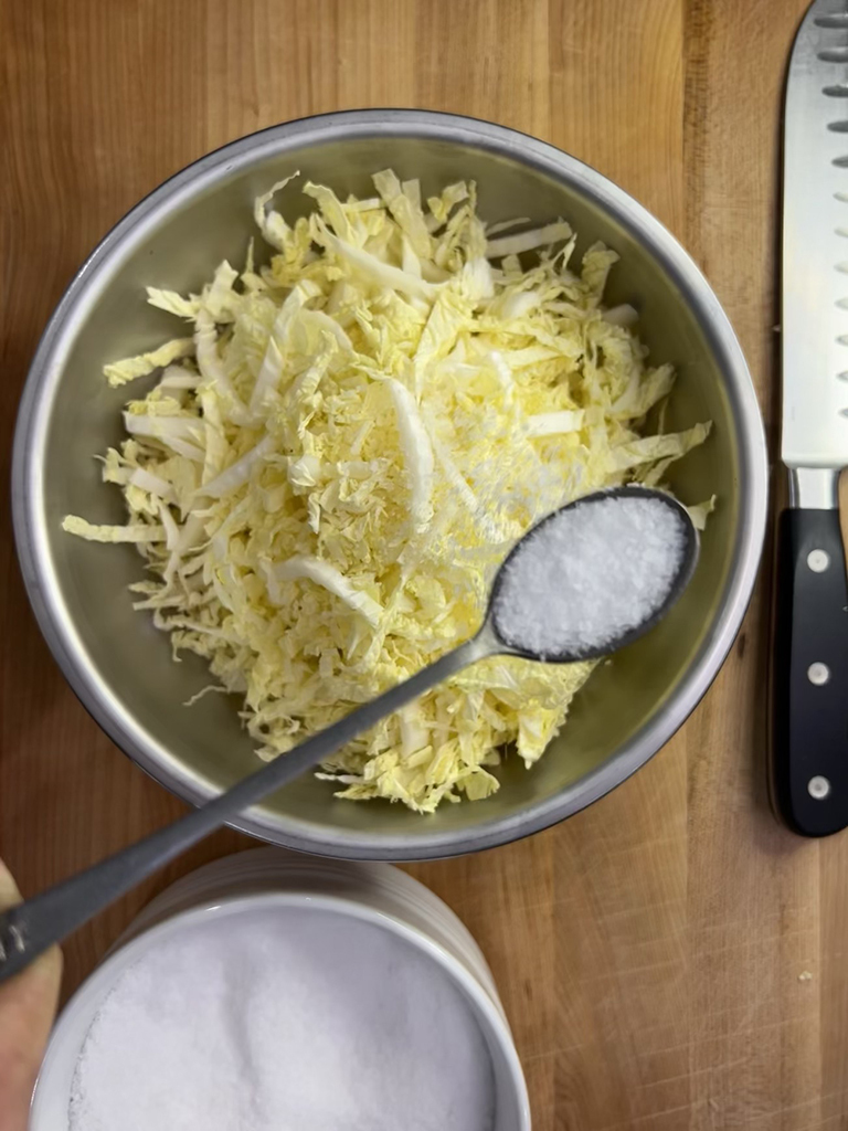A spoonful of salt hovers over a stainless steel bowl of finely sliced cabbage.