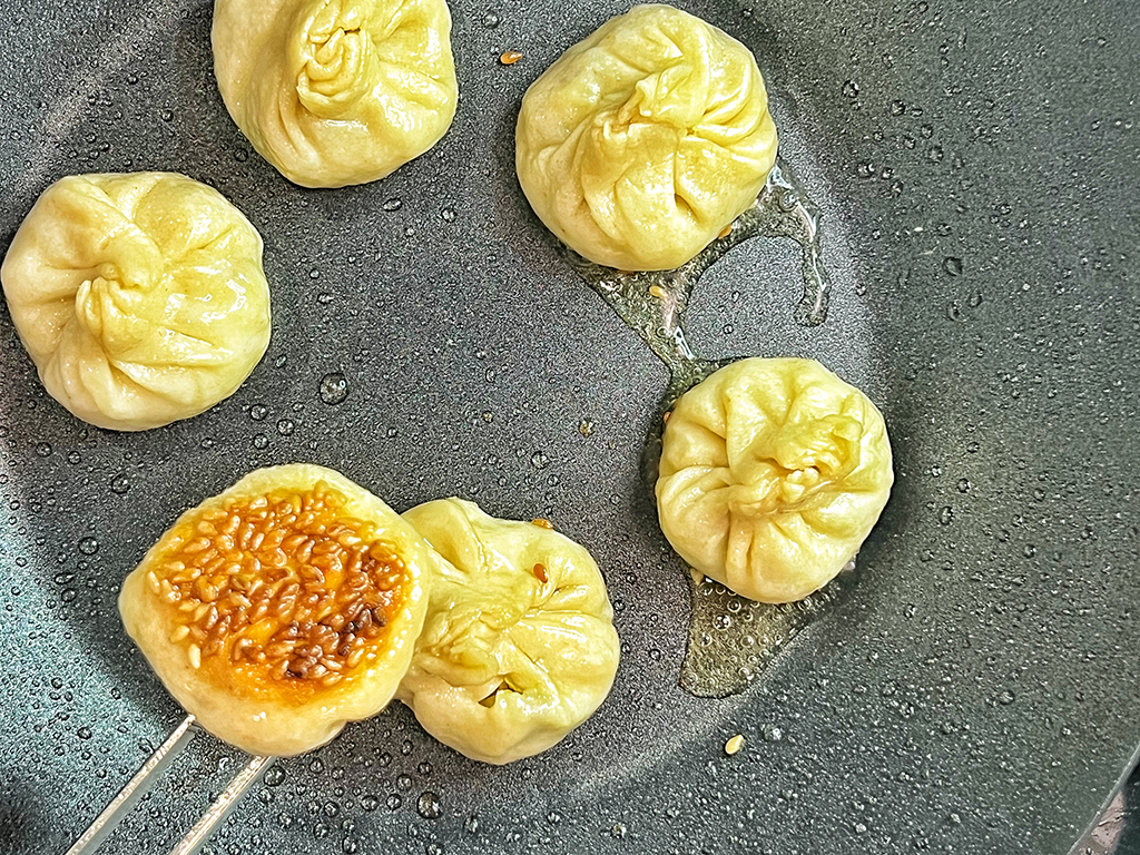 Six dumplings in a pan, frying in oil. Chopsticks hold one upside down, revealing a sesame seed crust.