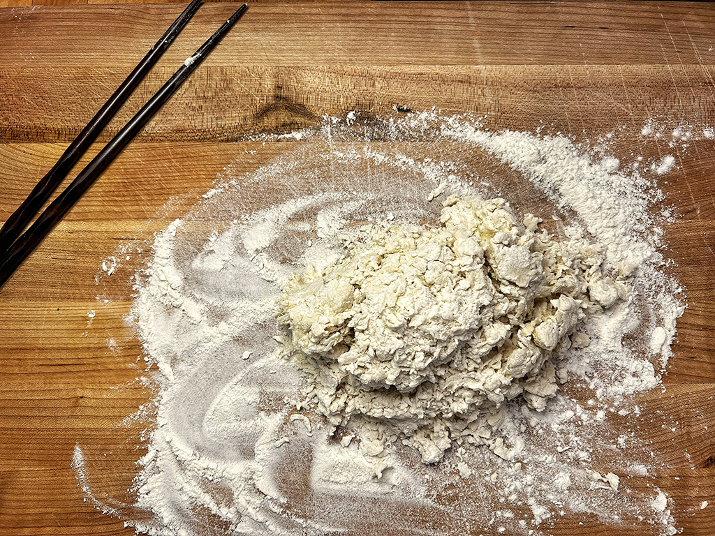 A raggy blob of dough on a floured countertop next to a pair of chopsticks.