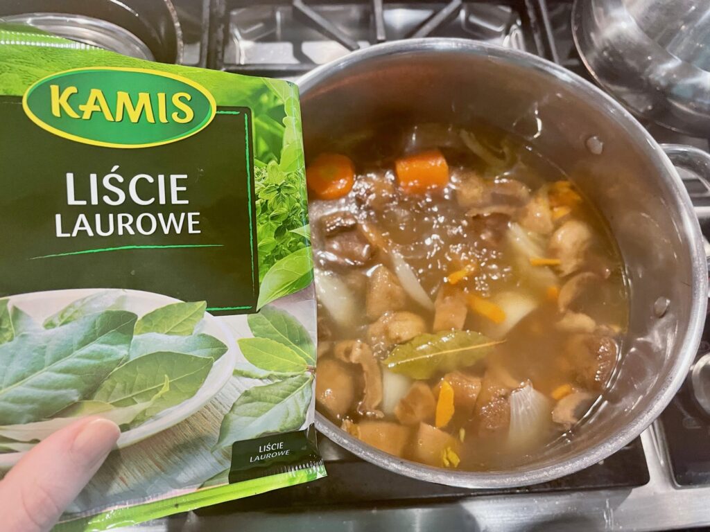 A hand holds a plastic back with a picture of bay leaves on it over a soup pot full of vegetables and one bay leaf. The label on the package is in Polish.