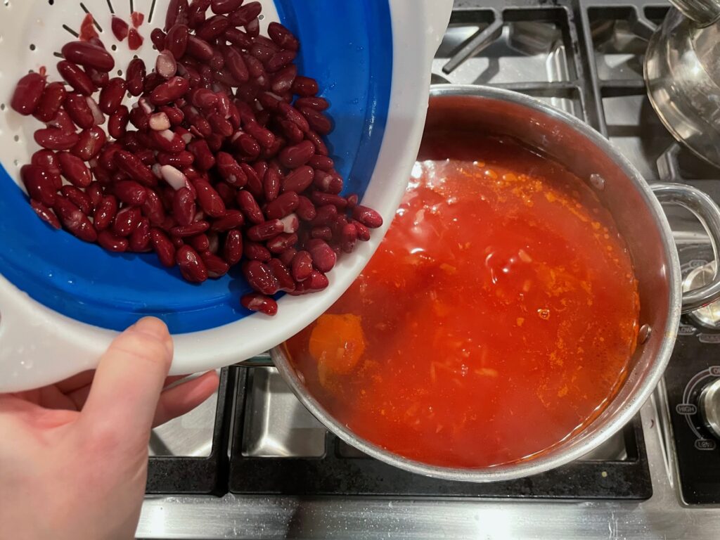 A hand holding a colander full of red kidney beans pours the beans into a pot of red soup on a stovetop.