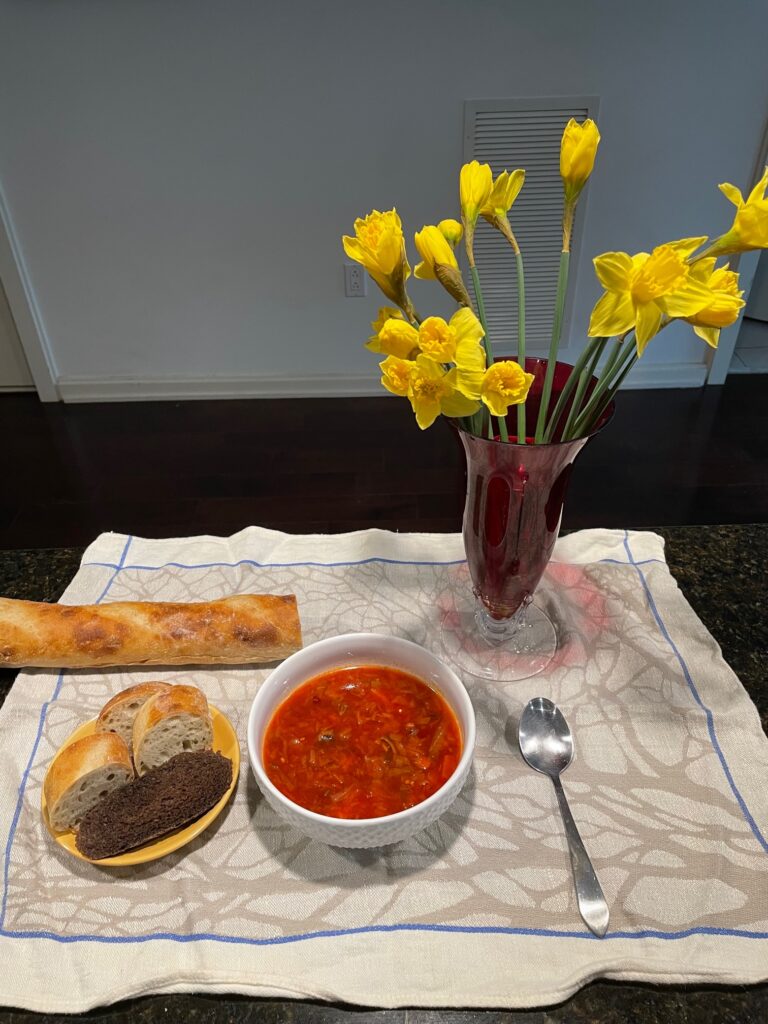 A bowl of borscht sits on a placemat. To its left, there is a plate with white and brown bread, as well as a baguette. To its right, there is a vase of daffodils and a spoon.