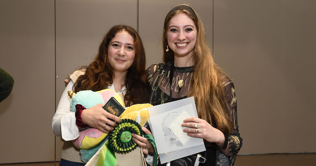 Elena and Katherine smile at the Edible Books Festival awards ceremony, holding armfuls of prizes include an enormous green prize ribbon.