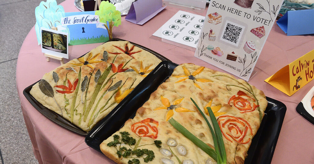Focaccia bread decorated with floral designs made out of vegetables sits on a table. Near it there is a small sign that says "The Secret Garden" and a second sign that says "Scan Now to Vote."
