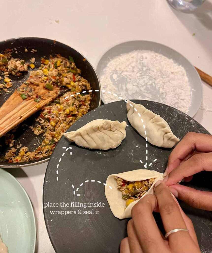 A frying pan with sauteed veggies and tofu sit in a pan next to a shallow dish of flour. On a third dish, two sealed dumplings sit next to a dumpling that is in the process of being sealed by a person's hands. The veggie filling is visible on the unsealed dumpling. A caption says, "place the filling inside the wrappers & seal it"
