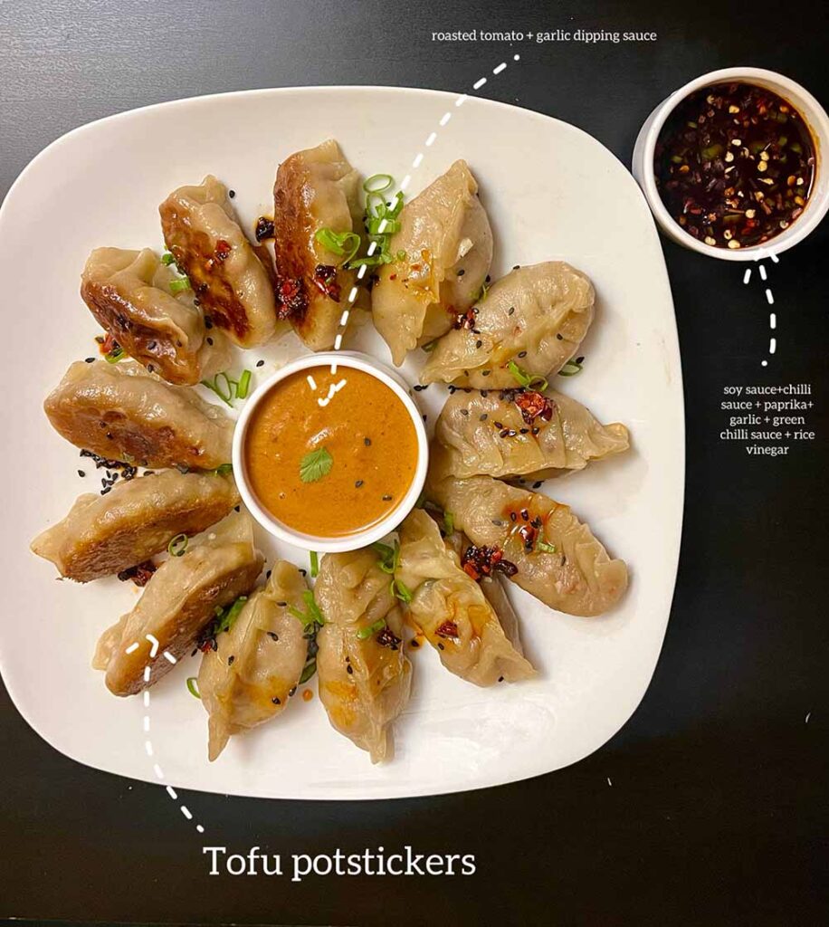 13 beautiful dumplings sit on a white platter, surrounding a cup of orange sauce. To the side of the platter is another cup filled with a dark brown sauce. A caption says "Tofu potstickers" with an arrow pointing at the dumplings. A caption says "roasted tomato + garlic dipping sauce" with an arrow pointing at the orange sauce. A caption says "Soy sauce and chili sauce + paprika + garlic + green chili sauce + rice vinegar" with an arrow pointing towards the brown sauce.